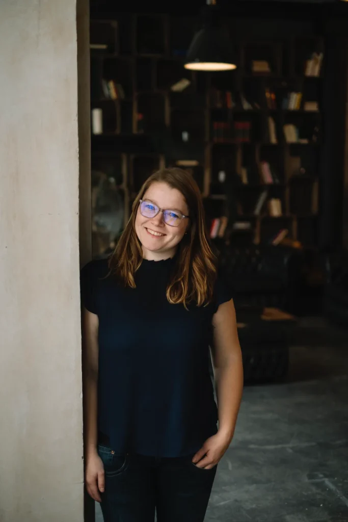 Junge Frau mit Brille steht neben einer Wand in einem Raum mit Bücherregalen im Hintergrund.