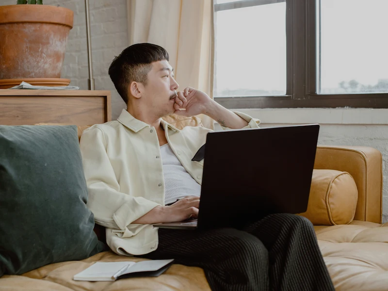 Mann sitzt auf einem Sofa und schaut nachdenklich aus dem Fenster, während er einen Laptop benutzt.