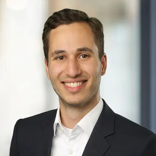 Porträt eines Mannes mit braunen Haaren und einem Lächeln, gekleidet in einem dunklen Anzug und einem weißen Hemd.