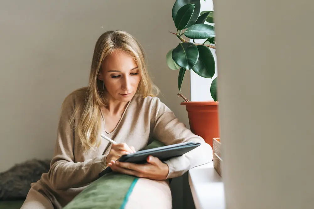 Eine blonde Frau schreibt konzentriert mit einem Stift auf einem Tablet, während sie neben einer großen Zimmerpflanze am Fenster sitzt.