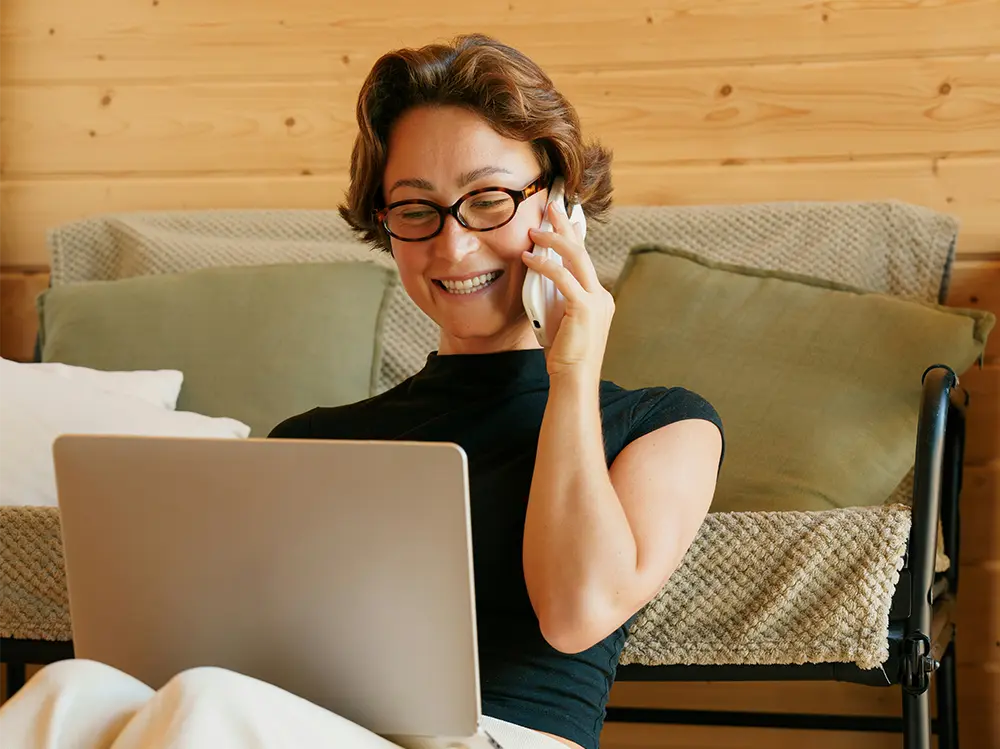 Mit Brille am Laptop telefoniert eine Frau lächelnd auf dem Sofa vor einer Holzvertäfelung und grünen Kissen in einem gemütlichen Raum.